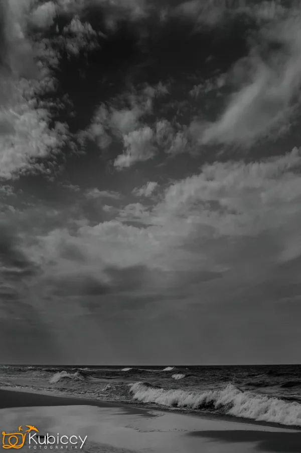 Czarno-białe zdjęcie plaży z falami, pod dramatycznym zachmurzonym niebem. Scena jest nastrojowa i klimatyczna. Logo Kubiccy jest widoczne w lewym dolnym rogu. - Kubiccy Fotografia - Fotograf Łódź