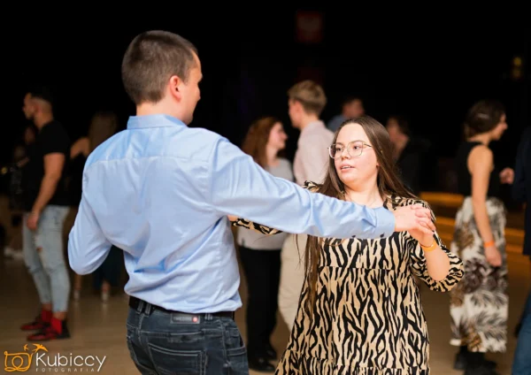 Mężczyzna w jasnoniebieskiej koszuli i kobieta w sukience z nadrukiem zebry tańczą razem w pomieszczeniu, otoczeni przez inne tańczące pary. Tło jest słabo oświetlone, a tancerze wydają się skupieni i szczęśliwi. - Kubiccy Fotografia - Fotograf Łódź