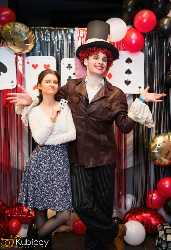 Dwie osoby w kostiumach pozują na przyjęciu z dekoracjami z kart do gry. Jedna z nich ma na sobie brązową kurtkę, cylinder i spodnie w paski, podczas gdy druga trzyma kartę do gry i ma na sobie kwiecistą spódnicę i białą bluzkę. Za nimi znajdują się balony i srebrne serpentyny. - Kubiccy Fotografia - Fotograf Łódź
