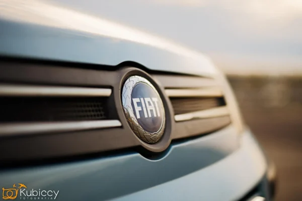 Zbliżenie na przedni grill i logo jasnoniebieskiego samochodu marki Fiat, z wyraźnie widocznym emblematem Fiata pośrodku. Tło jest rozmyte, skupiając uwagę na logo. - Kubiccy Fotografia - Fotograf Łódź