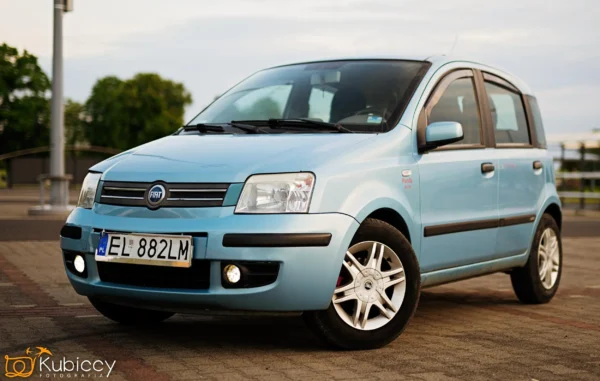 Jasnoniebieski samochód Fiat Panda hatchback jest zaparkowany na utwardzonym terenie na zewnątrz, z drzewami i zachmurzonym niebem w tle. Samochód ma polskie tablice rejestracyjne i felgi aluminiowe. - Kubiccy Fotografia - Fotograf Łódź