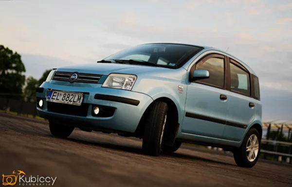 Jasnoniebieski samochód Fiat Panda zaparkowany na utwardzonej powierzchni o zachodzie słońca, widziany z niskiego kąta. Reflektory samochodu są włączone, a w tle widoczne jest zachmurzone niebo. Logo Kubiccy Fotografia znajduje się w lewym dolnym rogu. - Kubiccy Fotografia - Fotograf Łódź