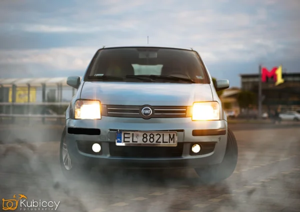 Srebrny samochód marki Fiat z włączonymi reflektorami jest zaparkowany na utwardzonej powierzchni, emitując dym z opon. Na tablicy rejestracyjnej widnieje napis EL 882LM. W tle widoczne jest centrum handlowe i zachmurzone niebo. - Kubiccy Fotografia - Fotograf Łódź