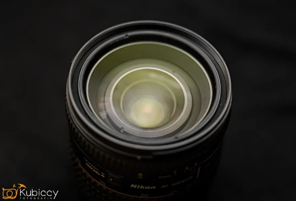 Zbliżenie obiektywu aparatu Nikon AF Nikkor z widocznymi szklanymi elementami, na ciemnym, rozmytym tle. W lewym dolnym rogu widnieje logo Kubiccy Fotografia. - Kubiccy Fotografia - Fotograf Łódź