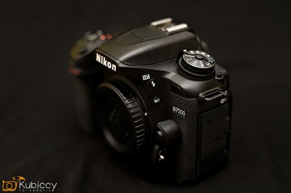 Zbliżenie czarnego korpusu lustrzanki cyfrowej Nikon D7500, umieszczonego na ciemnej powierzchni, z widocznymi przyciskami, pokrętłami i teksturowanym uchwytem. - Kubiccy Fotografia - Fotograf Łódź