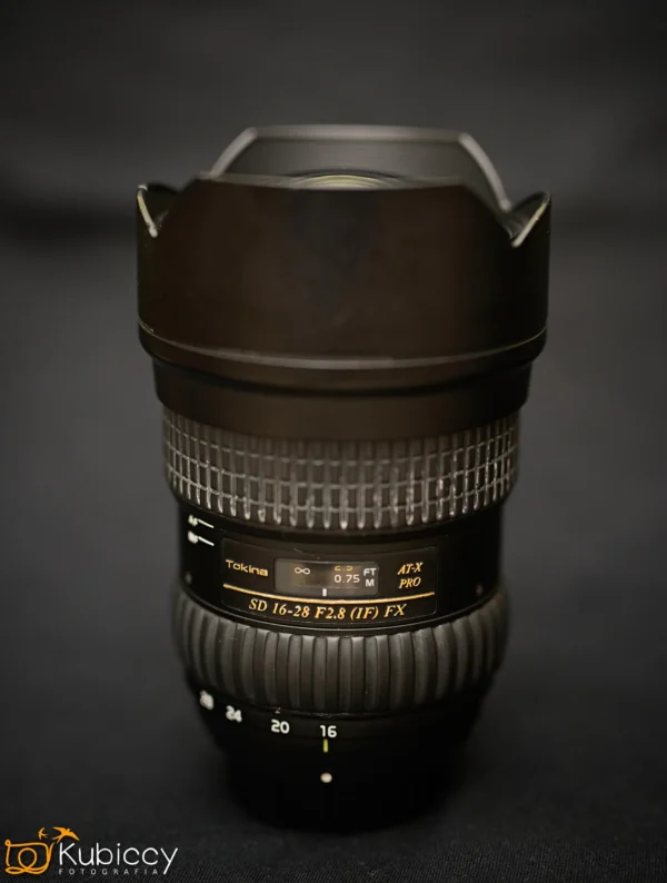 Czarny obiektyw Tokina AT-X PRO SD 16-28mm F2.8 (IF) FX stoi pionowo na ciemnej powierzchni, z widocznym logo i szczegółami obiektywu; tło jest czarne i rozmyte. - Kubiccy Fotografia - Fotograf Łódź