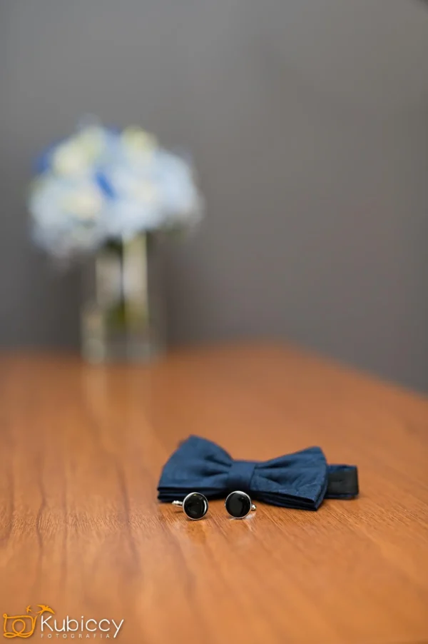 Granatowa muszka i para czarnych spinek do mankietów spoczywają na drewnianym stole, z rozmytym szklanym wazonem z niebieskimi i białymi kwiatami w tle. - Kubiccy Fotografia - Fotograf Łódź