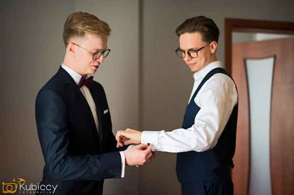 Dwóch mężczyzn w strojach wizytowych stoi w pomieszczeniu; jeden w garniturze pomaga drugiemu, który jest w kamizelce i koszuli, zapiąć spinki do mankietów. Obaj mają krótkie włosy, okulary i są skupieni na zadaniu. - Kubiccy Fotografia - Fotograf Łódź
