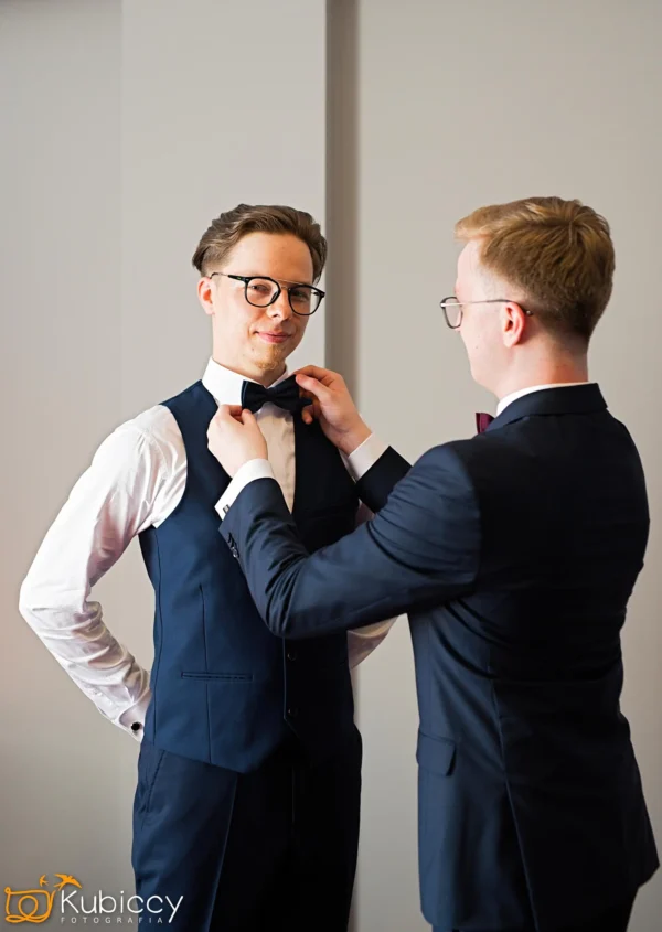 Dwóch mężczyzn w formalnych strojach stoi w pomieszczeniu; jeden reguluje muszkę drugiego. Obaj noszą okulary i granatowe kamizelki. Scena wydaje się lekka i wspierająca, wskazując na przygotowania do formalnego wydarzenia. - Kubiccy Fotografia - Fotograf Łódź