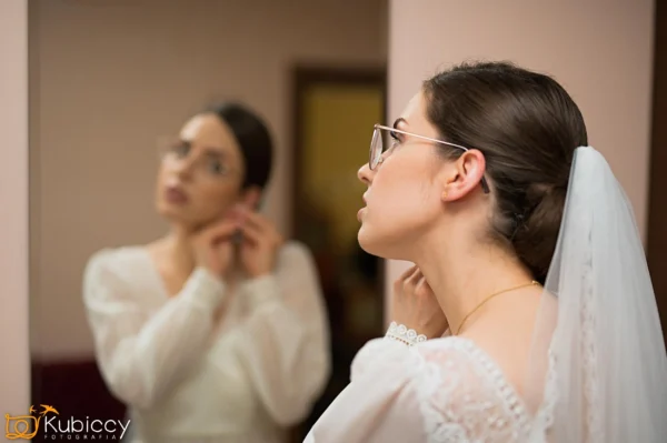 Panna młoda w okularach i białej sukni poprawia kolczyk, przeglądając się w lustrze, z włosami zaczesanymi do tyłu i welonem. - Kubiccy Fotografia - Fotograf Łódź