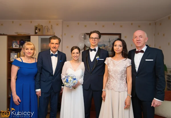 Para młoda stoi w centrum, uśmiechnięta, otoczona dobrze ubranymi rodzicami w ciepło oświetlonym pokoju ozdobionym balonami, kwiatami i półką z książkami w tle. - Kubiccy Fotografia - Fotograf Łódź