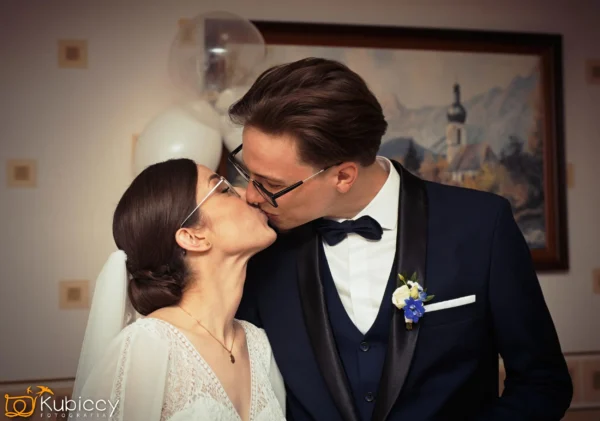Panna młoda i pan młody całują się w pomieszczeniu. Pan młody ma na sobie ciemny garnitur z muszką i butonierką, a panna młoda ma na sobie białą sukienkę i okulary. W tle widoczny jest obraz i balony. - Kubiccy Fotografia - Fotograf Łódź