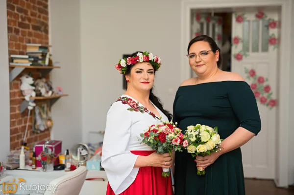 Dwie kobiety stoją w pomieszczeniu trzymając bukiety kwiatów. Jedna z nich ma na sobie kwiecistą koronę, białą bluzkę i czerwoną spódnicę; druga ma na sobie ciemnozieloną sukienkę z odkrytymi ramionami. W tle widoczna jest półka i otwarte drzwi. - Kubiccy Fotografia - Fotograf Łódź