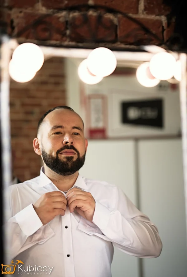 Brodaty mężczyzna w białej koszuli poprawia kołnierzyk, stojąc przed dobrze oświetlonym lustrem z odsłoniętymi żarówkami, przygotowując się. - Kubiccy Fotografia - Fotograf Łódź