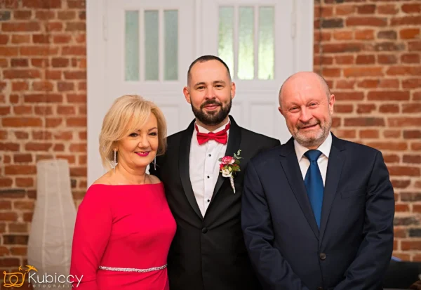 Trzy dorosłe osoby stoją uśmiechnięte w pomieszczeniu przed ceglaną ścianą i białymi drzwiami. Kobieta po lewej stronie ma na sobie czerwoną sukienkę, mężczyzna w środku ma na sobie smoking, a mężczyzna po prawej stronie ma na sobie garnitur. - Kubiccy Fotografia - Fotograf Łódź