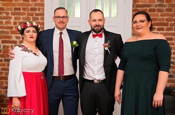 Cztery dorosłe osoby stoją blisko siebie w pomieszczeniu, ubrane formalnie. Dwaj mężczyźni noszą garnitury - jeden z czerwoną muszką i butonierką. Dwie kobiety noszą sukienki, jedną czerwoną z kwiatowym haftem, a drugą ciemnozieloną. Uśmiechają się przed ceglaną ścianą. - Kubiccy Fotografia - Fotograf Łódź