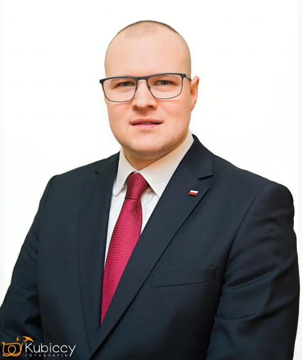 Mężczyzna z krótkimi włosami, w okularach, ciemnym garniturze, białej koszuli i czerwonym krawacie stoi na białym tle. Na klapie marynarki ma małą przypinkę z flagą Polski. Obraz posiada znak wodny Kubiccy Fotografia. - Kubiccy Fotografia - Fotograf Łódź