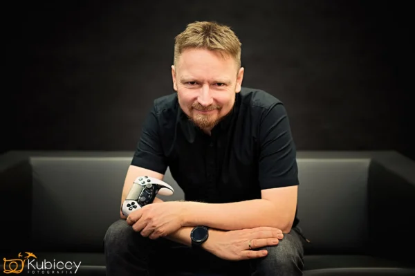 Uśmiechnięty mężczyzna z krótkimi włosami i brodą siedzi na ciemnej kanapie, pochylając się do przodu i trzymając kontroler gier wideo. Ma na sobie czarną koszulę i smartwatch, z rozmytym ciemnym tłem. - Kubiccy Fotografia - Fotograf Łódź