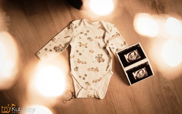 Dziecięca koszulka ze zwierzęcymi nadrukami leży na drewnianej podłodze obok czarno-białych zdjęć USG, otoczonych miękkimi, nieostrymi światłami. - Kubiccy Fotografia - Fotograf Łódź