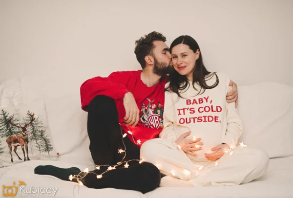 Para siedzi na białej kanapie, owinięta sznurkiem świateł. Mężczyzna całuje kobietę w policzek. Kobieta ma na sobie sweter z napisem "BABY, IT'S COLD OUTSIDE" i trzyma się za ciążowy brzuch. Obok nich leży poduszka z reniferem. - Kubiccy Fotografia - Fotograf Łódź