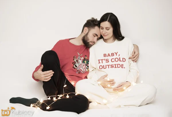 Para siedzi blisko siebie na białej kanapie. Kobieta, ubrana w kremową bluzę z napisem BABY, ITS COLD OUTSIDE, trzyma ciężarny brzuch. Mężczyzna opiera głowę na jej ramieniu. Wokół nich rozwieszone są światła. - Kubiccy Fotografia - Fotograf Łódź
