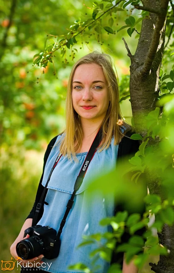 Młoda kobieta o blond włosach stoi na zewnątrz przy drzewie, ubrana w niebieski top i czarny sweter. Na szyi ma aparat fotograficzny i delikatnie się uśmiecha. Otaczają ją bujne zielone liście i rozmyte pomarańczowe jagody. - Kubiccy Fotografia - Fotograf Łódź