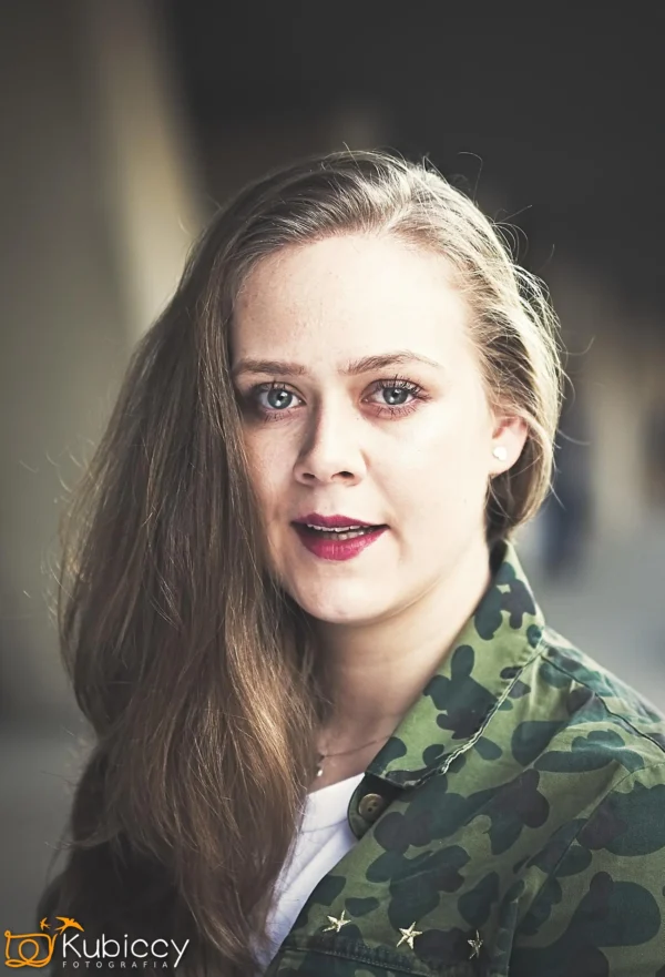 Kobieta z długimi jasnobrązowymi włosami, ubrana w zieloną kamuflażową kurtkę i białą koszulę, patrzy bezpośrednio w kamerę z neutralnym wyrazem twarzy. Tło jest rozmyte. - Kubiccy Fotografia - Fotograf Łódź
