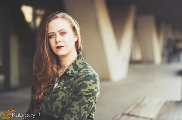 Kobieta z długimi włosami ubrana w kamuflaż siedzi na zewnątrz na drewnianej ławce, patrząc w kamerę. Tło jest rozmyte, ukazując betonowe kolumny i chodnik. - Kubiccy Fotografia - Fotograf Łódź