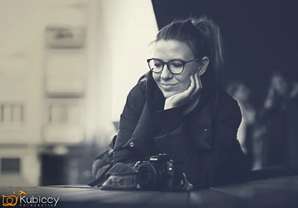 Kobieta w okularach uśmiecha się, opierając podbródek na dłoni. Ma na sobie kurtkę, a przed sobą kamerę. Obraz jest czarno-biały. - Kubiccy Fotografia - Fotograf Łódź