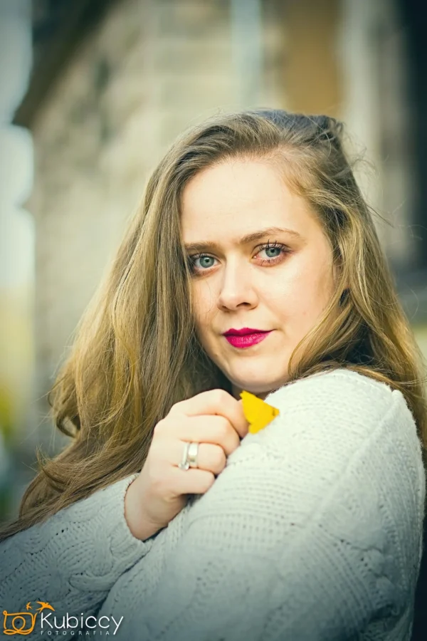 Kobieta z długimi blond włosami i jasną szminką ma na sobie jasny sweter, a przy twarzy trzyma mały żółty liść. Patrzy bezpośrednio w kamerę z łagodnym, neutralnym wyrazem twarzy. - Kubiccy Fotografia - Fotograf Łódź