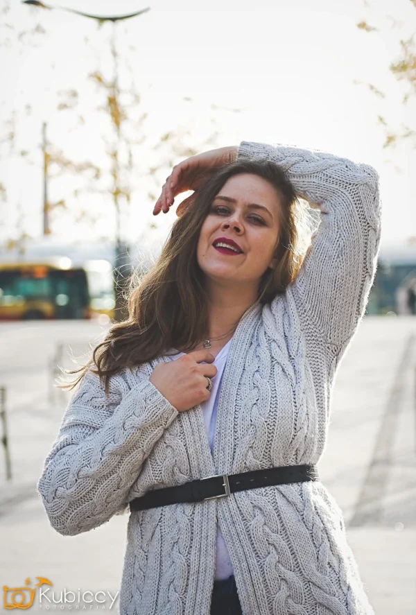Kobieta z długimi brązowymi włosami ubrana w szary dzianinowy kardigan i czarny pasek pozuje na zewnątrz, uśmiechając się z jedną ręką uniesioną nad głową. Drzewa, autobus i ulica są widoczne w rozmytym tle. - Kubiccy Fotografia - Fotograf Łódź