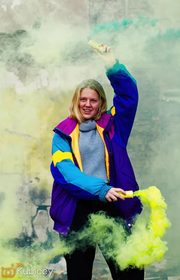 Osoba w kolorowej kurtce trzyma żółte race dymne, otoczone gęstym żółtym i zielonym dymem, stojąc przed zwietrzałą ścianą. - Kubiccy Fotografia - Fotograf Łódź