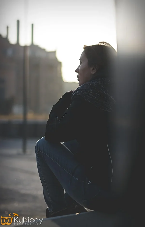 Osoba siedzi na zewnątrz na krawężniku o zachodzie słońca, opierając ręce na kolanach i patrząc w dal. Scena jest delikatnie oświetlona, tworząc kontemplacyjny nastrój. W rogu widoczne jest logo Kubiccy Fotografia. - Kubiccy Fotografia - Fotograf Łódź