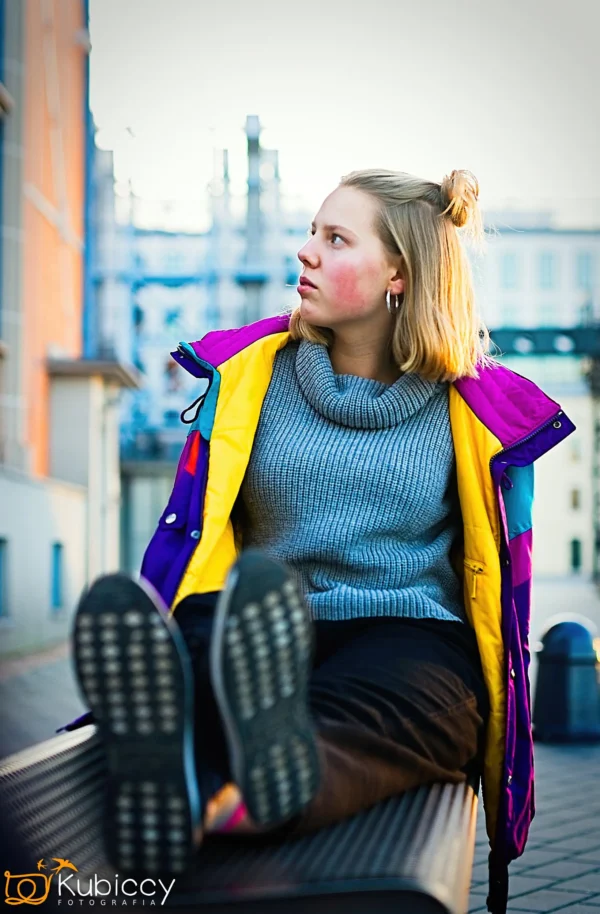 Młoda kobieta w kolorowej kurtce i szarym swetrze siedzi na ławce z nogami w górze, patrząc w bok. W tle widać miejskie budynki i konstrukcje przemysłowe. - Kubiccy Fotografia - Fotograf Łódź