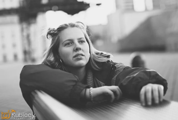 Młoda kobieta w jasnych włosach i kurtce opiera się o metalową barierkę na zewnątrz, patrząc w zamyśleniu w dal. Zdjęcie jest czarno-białe z rozmytym miejskim tłem. - Kubiccy Fotografia - Fotograf Łódź