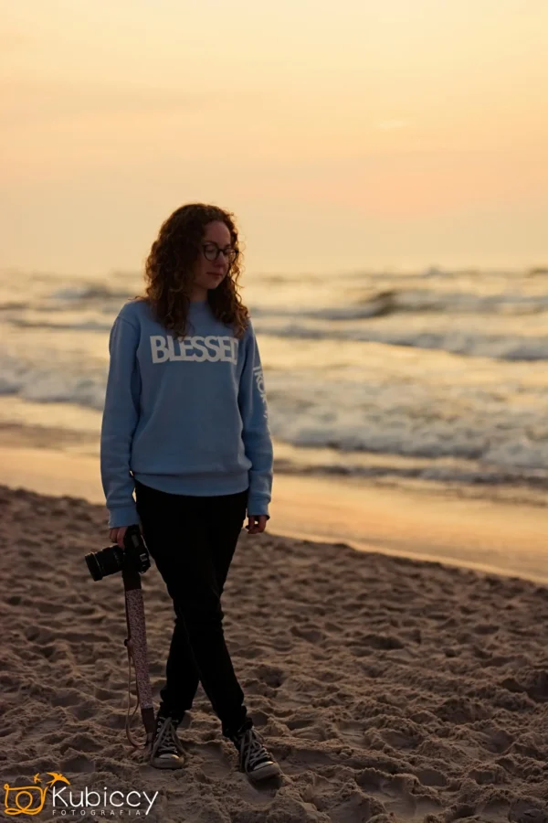 Kobieta z kręconymi włosami, w okularach i jasnoniebieskiej bluzie BLESSED idzie boso po piaszczystej plaży o zachodzie słońca, trzymając aparat w prawej ręce. W tle widoczne są fale i ciepłe niebo. - Kubiccy Fotografia - Fotograf Łódź