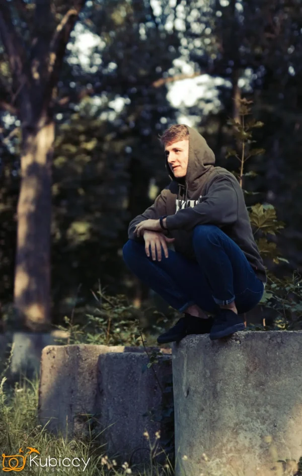 Osoba w bluzie z kapturem i dżinsach kuca na betonowym bloku na zewnątrz, otoczona zielenią i drzewami, a światło słoneczne filtruje przez tło. - Kubiccy Fotografia - Fotograf Łódź