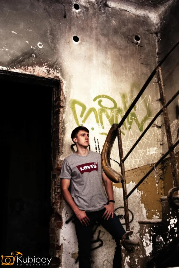 Młody mężczyzna w szarej koszulce Levi's stoi na zniszczonych schodach w opuszczonym budynku z pokrytymi graffiti, łuszczącymi się ścianami. Ostre oświetlenie tworzy dramatyczną, miejską atmosferę. - Kubiccy Fotografia - Fotograf Łódź