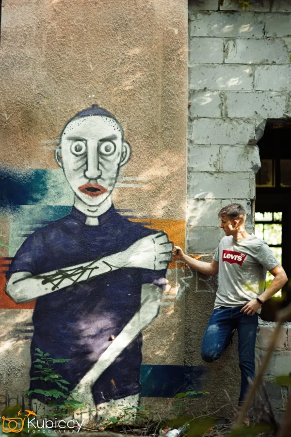 Mężczyzna w szarej koszulce Levi's pozuje przy muralu przedstawiającym wytatuowaną postać o bladej skórze i intensywnych oczach na zwietrzałej ścianie, obok częściowo zrujnowanego budynku i zarośniętych roślin. - Kubiccy Fotografia - Fotograf Łódź