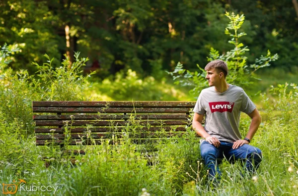 Młody mężczyzna w szarej koszulce Levi's i dżinsach siedzi na zwietrzałej drewnianej ławce w trawiastym, zarośniętym parku, patrząc w prawo. Światło słoneczne przenika przez bujne zielone drzewa w tle. - Kubiccy Fotografia - Fotograf Łódź