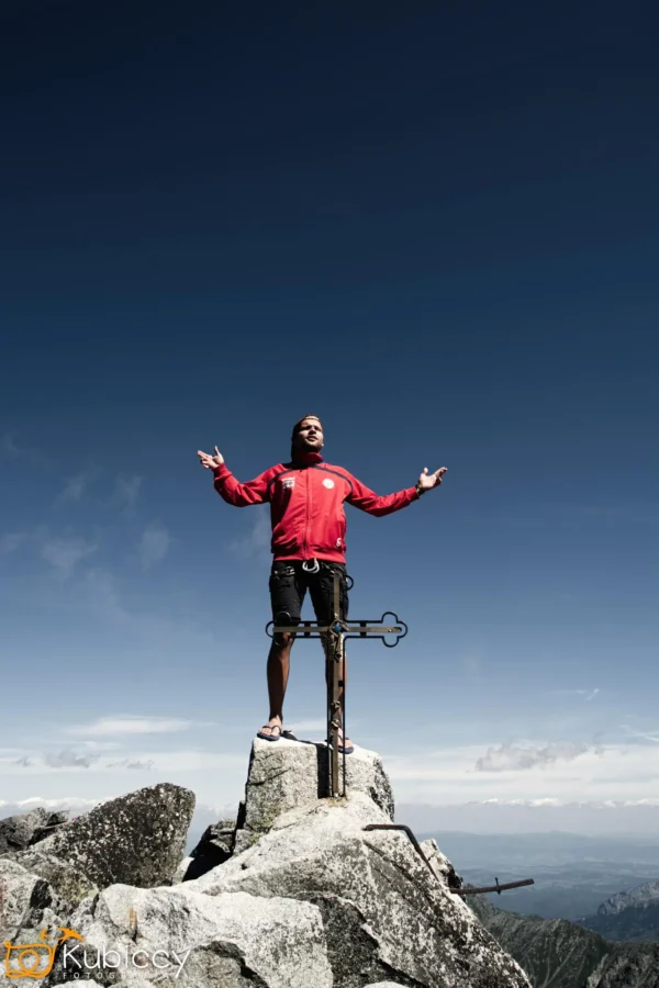 Osoba w czerwonej kurtce stoi triumfalnie na skalistym szczycie góry, z wyciągniętymi ramionami, z metalowym znacznikiem szczytu obok i czystym, błękitnym niebem w tle. - Kubiccy Fotografia - Fotograf Łódź