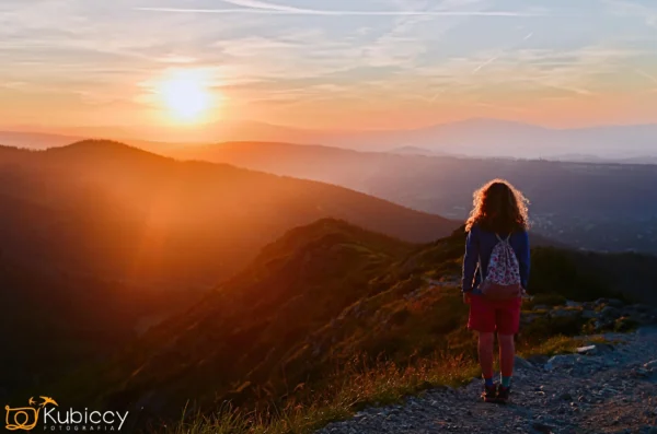 Młoda dziewczyna z kręconymi włosami stoi na górskiej ścieżce o zachodzie słońca, z widokiem na falujące wzgórza i doliny skąpane w złotym słońcu. Niebo jest usiane chmurami i smugami. - Kubiccy Fotografia - Fotograf Łódź