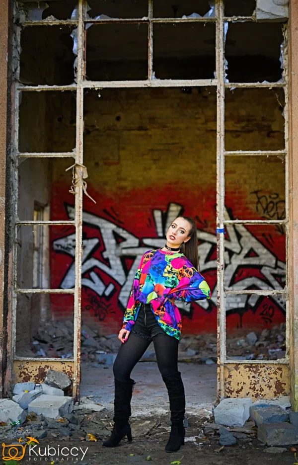 Kobieta w kolorowej bluzce i czarnych butach pozuje pewnie przed rozbitą ramą okna, z graffiti i rozpadającymi się cegłami widocznymi w opuszczonym budynku za nią. - Kubiccy Fotografia - Fotograf Łódź