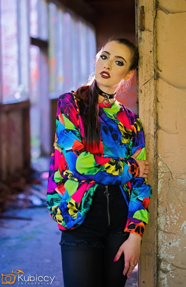 Młoda kobieta z długimi brązowymi włosami spiętymi w kucyk opiera się o zniszczone drzwi. Ma na sobie kolorowy sweter w abstrakcyjne wzory, czarne szorty i odważny makijaż z ciemną szminką i brokatem nad brwiami. - Kubiccy Fotografia - Fotograf Łódź