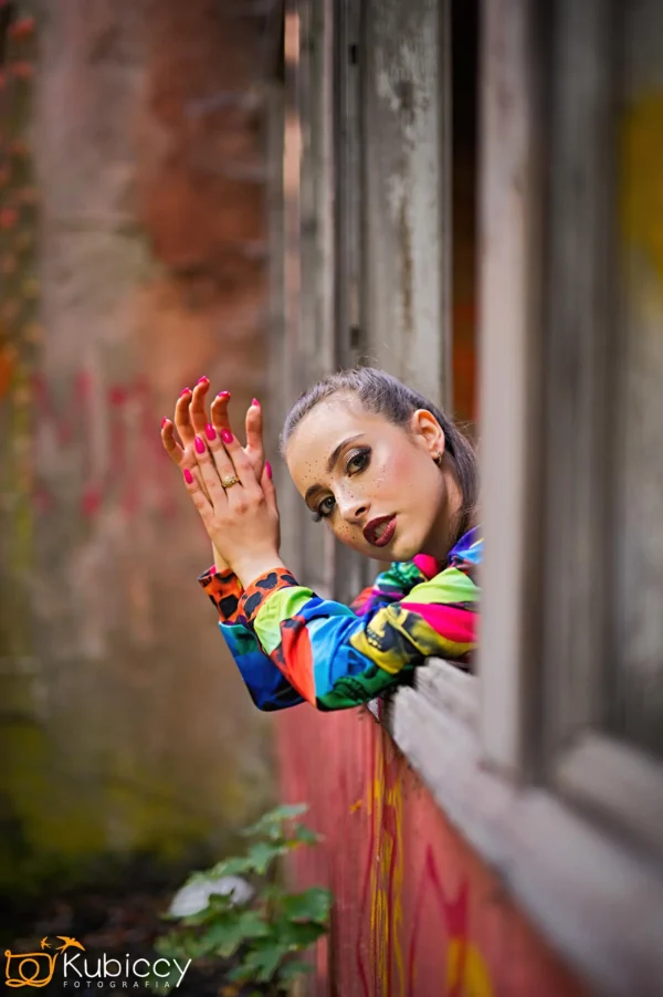 Kobieta z wyrazistym makijażem i kolorową kurtką wychyla się przez zwietrzałe okno, z podniesionymi rękami i splecionymi palcami, na tle starego, pokrytego graffiti budynku. - Kubiccy Fotografia - Fotograf Łódź