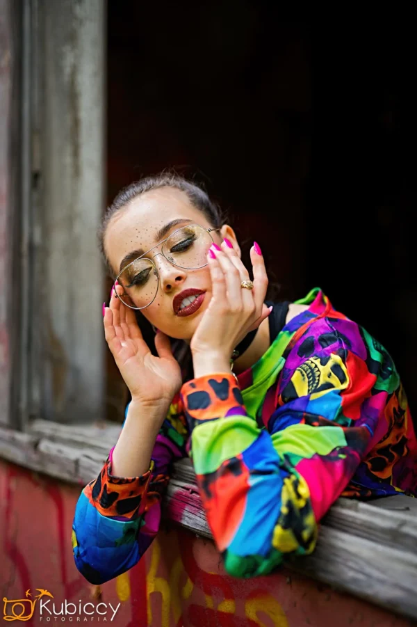 Osoba z jasnoróżowymi paznokciami i dużymi okrągłymi okularami opiera się o parapet, ubrana w kolorową, wzorzystą koszulę i wygląda na zrelaksowaną, z dłońmi delikatnie okalającymi twarz. - Kubiccy Fotografia - Fotograf Łódź