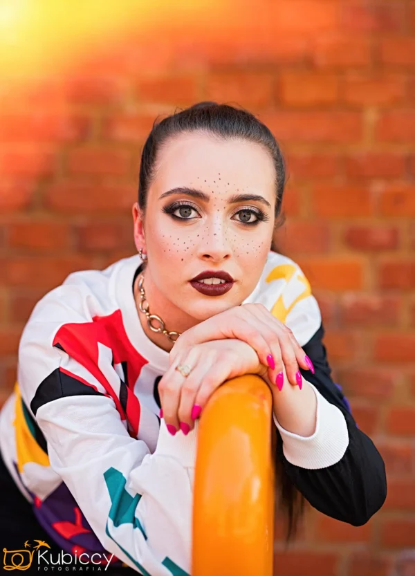 Kobieta z odważnym makijażem, w tym ciemną szminką i dramatycznym eyelinerem, opiera ręce na pomarańczowej poręczy. Ma na sobie kolorowy top i różowe paznokcie. W tle widać ceglaną ścianę. - Kubiccy Fotografia - Fotograf Łódź