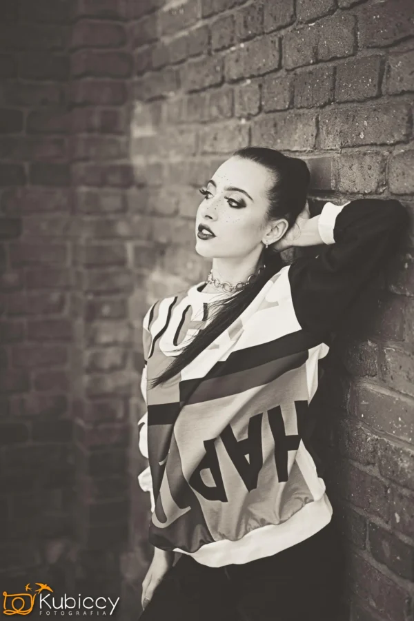 Kobieta z wyrazistym makijażem opiera się o ceglaną ścianę, spoglądając w górę. Ma na sobie bluzę z grafiką i włosy zaczesane do tyłu. Obraz jest czarno-biały, z logo w lewym dolnym rogu. - Kubiccy Fotografia - Fotograf Łódź