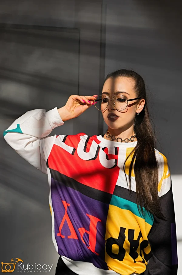 Kobieta w dużych okrągłych okularach, ciemnej szmince i kolorowej graficznej bluzie pozuje z jedną ręką na okularach. Światło słoneczne rzuca cienie na ścianę za nią. - Kubiccy Fotografia - Fotograf Łódź