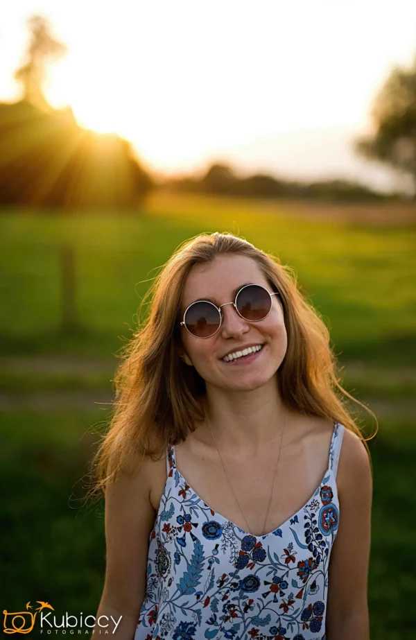 Młoda kobieta z długimi włosami w okrągłych okularach przeciwsłonecznych i kwiecistej sukience uśmiecha się na zewnątrz na oświetlonym słońcem polu, z zielenią i drzewami w tle. Zachodzące słońce tworzy za nią ciepłą poświatę. - Kubiccy Fotografia - Fotograf Łódź